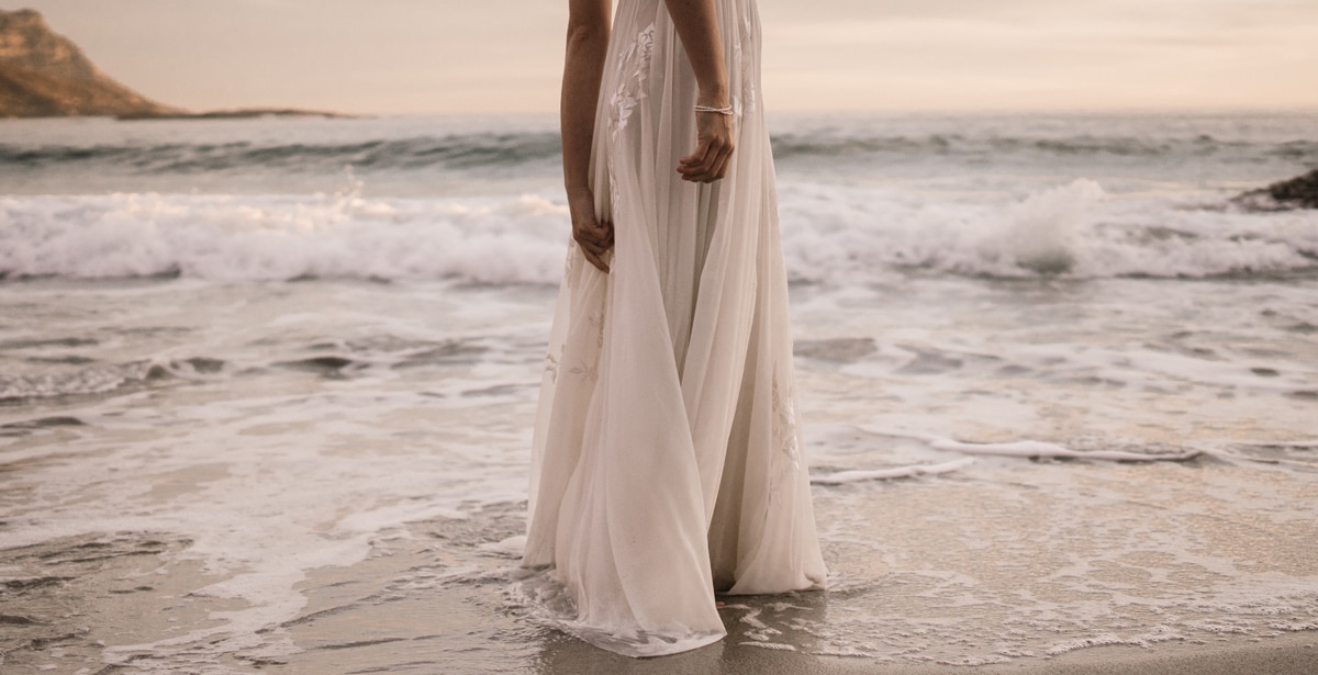 Bride stands on the beach by the water.